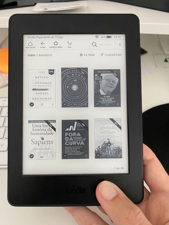 A imagem mostra o Kindle na tela de visualização da biblioteca pessoal de eBooks. No caso, aparecem 6 eBooks, que são apenas uma parte dessa biblioteca. Os 6 eBooks são: Rápido e Devagar, Para explicar o mundo, O jeito Peter Lynch de investir, Sapiens, Fora da Curva e A invenção da natureza.